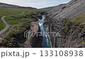 Aerial view of Studlagil Canyon in Iceland, showcasing basalt columns, a turquoise river, steep cliffs, greenery, and small waterfalls in motion. 133108938