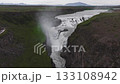 Aerial view of Gullfoss waterfall in Iceland, with cascading water, mist, walking paths, green vegetation, rocky terrain, and distant mountains. 133108942