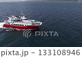 A red and white boat moves steadily across deep blue ocean waters, leaving a white wake. Snow capped mountains and a rugged coastline are visible. 133108946