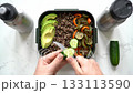 Top-down view of hands slicing cucumber near a wellness lunchbox with quinoa, avocado, and veggies on kitchen counter. 133113590