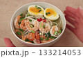 Asian dish with broth, noodles, tiger shrimp, fish, egg and green onions. The woman turns the plate. Traditional Asian soup in a white deep plate. Selective focus, close-up. 133214053