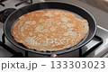 A golden-brown pancake cooking in a black frying pan on a gas stove. The pancake has a smooth texture and is evenly cooked. 133303023