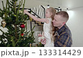 Father holding young daughter while decorating christmas tree, hanging ornaments together in warmly lit home, sharing festive family moment 133314429