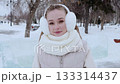 Blonde woman wearing winter accessories standing in snowy park, warming hands with gloved fingers and radiating cheerful smile 133314437