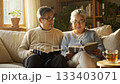Senior Asian couple sitting on a sofa holding books and smiling at each other in a sunlit room. This cozy scene depicts love in retirement, suitable for lifestyle and family content. 133403071