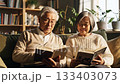 Happy senior Asian couple reading books together on a sofa in a sunlit living room. They look at each other with a smile, enjoying retirement and leisure time at home. 133403073