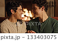 Two happy men sit close by burning fireplace looking into each other's eyes. Intimate moment represents LGBTQ love and domestic comfort. Ideal for relationship concepts. 133403075