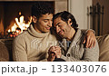Two affectionate men sit on sofa holding hands near burning fireplace. The cozy romantic scene conveys intimacy and peace. Ideal for relationship and lifestyle concepts. 133403076