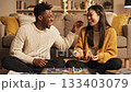 Cheerful interracial couple sits on floor playing board game. Cozy evening atmosphere with laughter and fun. Perfect for lifestyle, relationship and leisure concepts. 133403079