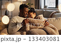Happy multiethnic couple cuddles under knitted blanket on sofa in dim living room. Scene evokes warmth, intimacy and relaxation. Perfect for lifestyle and relationship themes. 133403081