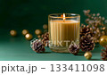 A glowing candle in a glass holder surrounded by pine cones and golden decor on a soft fabric, creating a warm festive atmosphere for winter celebration 133411098