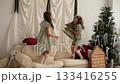 The girls are playing with a green Christmas tree-shaped pillow, jumping merrily on the couch and throwing the toy to each other. New Year at home 133416255