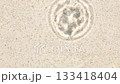 Water droplet impacts sandy surface, forming symmetrical ripple waves. Sunlight enhances translucent rings in fluid motion. Minimalist scene of natural rhythm and energy. 133418404