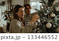 Young smiling couple in cozy sweaters decorating Christmas tree with white ornament in bright modern home interior 133446552
