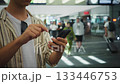 Young man views his smartphone, Man in striped shirt uses phone amid busy terminal scene, Young individual in casual attire interacts with mobile device amidst bustling transportation hub 133446753