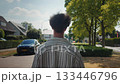 Young man walking across busy street from sidewalk, rear view shows leafy trees and oncoming cars, contemplative moment in neighborhood with warm light and steady composition 133446796