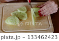 Woman with red nails slicing cucumber on wooden board, food preparation 133462423