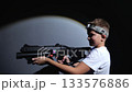 A happy teenage boy with a laser pistol is having fun in a dark laser tag arena, he is running around the arena and looking for a teammate. A colorful laser tag arena with neon stripes on the walls. 133576886