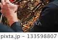 Close-up of a man playing a French horn. The hands of a musician clutching a shiny brass French horn, showing intricate valves and tubes, against a blurred background of fellow musicians 133598807