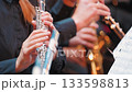 A close-up of musicians playing wind instruments during a performance. Focused on a flutist's hands, with blurred saxophones in the background, capturing the harmony of the ensemble 133598813