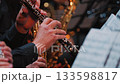 A close-up of musicians' hands playing woodwind instruments, specifically an oboe and a flute. The warm background features blurred brass instruments and sheet music, creating an musical atmosphere 133598817