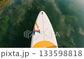 A view from above a paddle board gliding over calm, clear waters reflecting clouds and greenery below. The paddle board displays colorful markings and intricate details 133598818