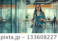Smiling businesswoman confidently walking through a modern office corridor, holding a folder, representing success, career advancement, and a positive professional journey 133608227