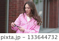 A young woman in a pink shirt looks at her smartphone while walking outside. The scene reflects modern digital lifestyle, showing how technology connects but also consumes attention daily. 133644732