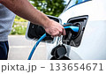 Close-up of man’s hand plugging in electric vehicle charger. Shiny blue car surface reflects ambient light. Concept of clean energy, EV technology, green transportation. Modern detail. Banner. 133654671