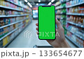 Hand holding smartphone with green screen in supermarket aisle. Blurred shelves with colorful products. Perfect mockup for app, e-commerce, or retail design. Bright and clean background. Banner 133654677