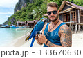 Man with tattoo poses on the beach holding a blue macaw, surrounded by tropical scenery and wooden huts at sunny seaside 133670630
