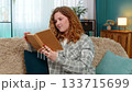 Woman sitting on sofa opening and reading book with focus thoughtfully flipping pages and reflecting 133715699