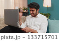 Indian man excited, celebrating project success, reacting with joy, smiling at laptop screen on sofa 133715701
