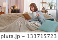 Smiling young woman reading interesting book while having cup of hot chocolate on couch at home 133715723