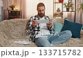 Young man sorting bills at home, sofa budget check, phone in hand, multiple papers, calm focus vibe 133715782