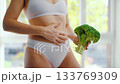 Woman is holding a broccoli in her hand. She strokes and squeezes her stomach 133769309