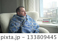 A man sits comfortably in a chair, wrapped in a warm blanket, gazing out of a window in his apartment. The cloudy weather outside adds to the cozy atmosphere as he enjoys a quiet moment. 133809445