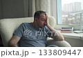 A man sits thoughtfully in a comfortable chair, gazing out the window. He appears contemplative, lost in his own thoughts. The urban skyline is visible in the background. 133809447