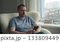 A man sits comfortably in a cozy chair by the window, focused on his phone. The city skyline is visible outside, creating a calm atmosphere for his afternoon. 133809449