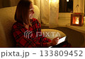 A woman relaxes on a comfy chair in a softly lit room. She enjoys her book, surrounded by candlelight, creating a peaceful, cozy atmosphere. 133809452