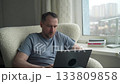 A man sits comfortably in a chair, focused on his laptop while enjoying a view of the city outside. The afternoon light creates a cozy atmosphere in his home. 133809858