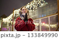 A young beautiful girl in a red coat walks in the evening at the European Christmas Market against the backdrop of an ice rink with illuminations and garlands. 133849990
