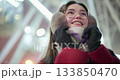 A young beautiful girl in a red coat walks in the evening at the New Year's Christmas fair against the backdrop of a Ferris wheel among lights and garlands and admires the decorations 133850470