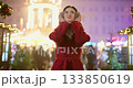 A young beautiful girl in a red coat walks in the evening at the European Christmas Market against the backdrop of illuminations and garlands. 133850619