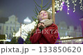 A young beautiful girl in a red coat walks in the evening at the European Christmas Market against the backdrop of an ice rink with illuminations and garlands. 133850620