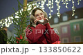 A young beautiful girl in a red coat walks in the evening at the European Christmas Market against the backdrop of an ice rink with illuminations and garlands. 133850622