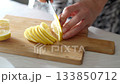 Someone carefully cuts a bright yellow lemon into thin slices on a wooden cutting board, preparing it for culinary use in a bright kitchen. 133850712
