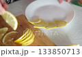 Close-up of hands artistically placing lemon slices around the rim of a white plate on a wooden cutting board, creating a beautiful citrus design in daylight. 133850713