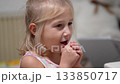 A cute child is shown seated. She carefully opens, eats, and savors a fruity vitamin supplement, expressing joy and satisfaction with each bite in the home. 133850717