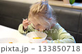 A small girl eats a bowl of soup in a calm Restaurant. The table is white and a sunny afternoon. She's enjoying her lunch and exploring flavors. 133850718
